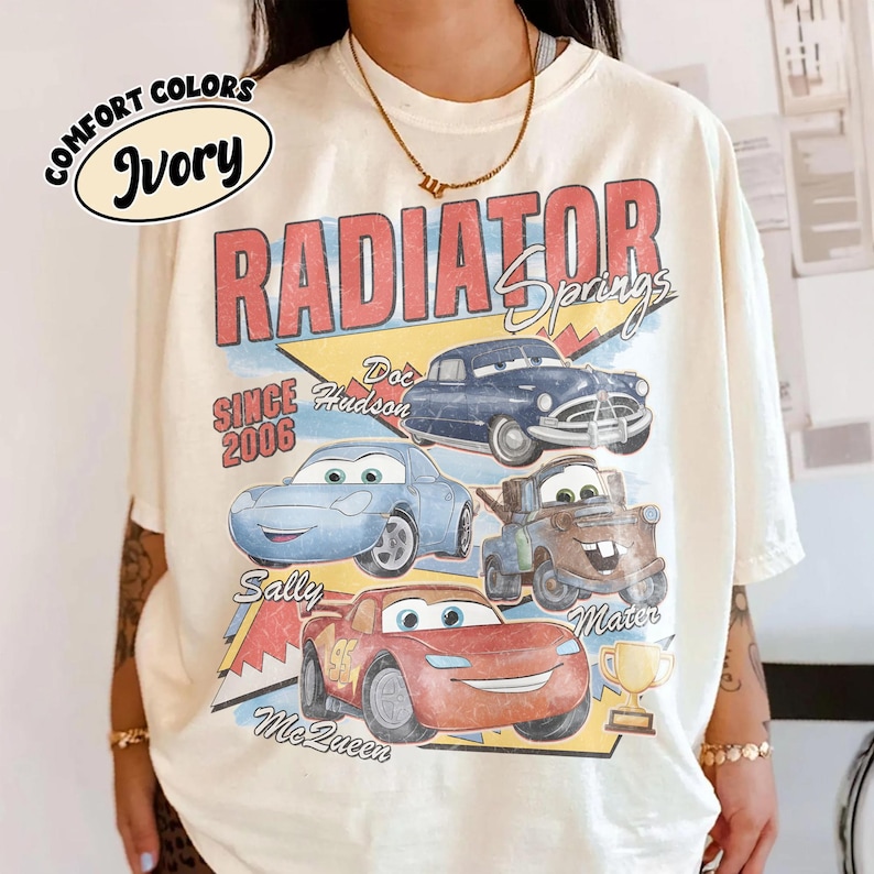 May include: Ivory-colored t-shirt featuring a graphic of characters from the movie Cars, including Doc Hudson, Sally, and Lightning McQueen. The design includes the text "Radiator Springs" and "Since 2006."