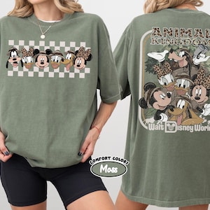K&ouml;nnte beinhalten: Olivgr&uuml;nes T-Shirt mit einer Grafik von Disney-Figuren, darunter Micky Maus, Minnie Maus, Donald Duck und Goofy, mit einem Schachbrettmuster. Auf der R&uuml;ckseite des Shirts stehen die Worte "Animal Kingdom" und "Walt Disney World".