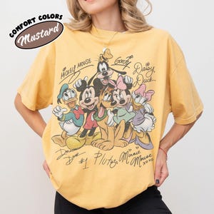 May include: Mustard-colored t-shirt featuring a graphic of Mickey Mouse, Donald Duck, Goofy, Pluto, Minnie Mouse, and Daisy Duck. The shirt has the text "Comfort Colors" and character names. The shirt is a relaxed fit.