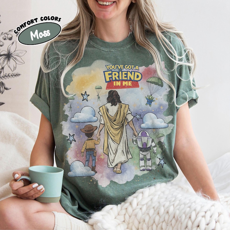 May include: Moss green t-shirt with a colorful graphic featuring the text "YOU'VE GOT A FRIEND IN ME." The design includes illustrations of Jesus, Woody, Buzz Lightyear, and other cartoon elements. The shirt has rolled-up sleeves.
