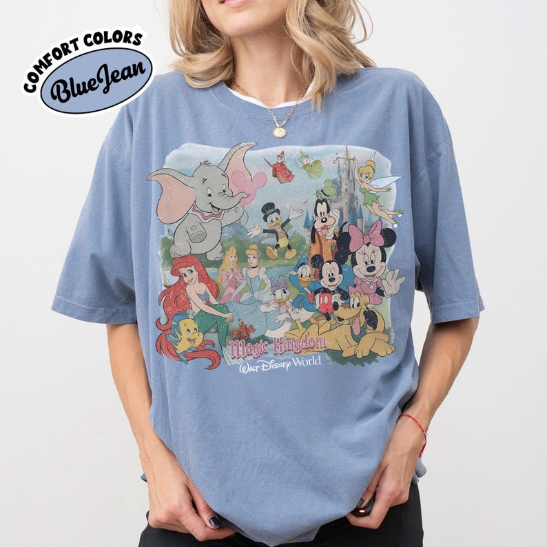 May include: Blue t-shirt with a graphic featuring Disney characters like Dumbo, Ariel, Mickey Mouse, and Minnie Mouse. The shirt has the text "Magic Kingdom Walt Disney World" and the words "Comfort Colors Blue Jean" in a circular design.