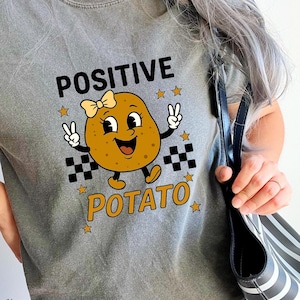 Puede incluir: Camiseta gris con un personaje de patata de dibujos animados con un lazo y haciendo el signo de la paz. El texto dice "POSITIVE POTATO" con acentos de estrella. Parte de la colección "The Chaos Collective".