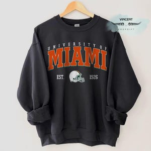 Pode incluir: Camisola preta de gola redonda com o texto "UNIVERSITY OF MIAMI" em letras laranja com contorno verde. Abaixo, um capacete de futebol americano branco e a inscrição "EST. 1926". A camisola está pendurada num cabide de madeira.