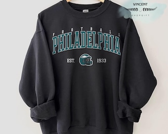 Può includere: Felpa nera con la scritta "FOOTBALL PHILADELPHIA" in lettere turchesi e bianche. Sotto, una grafica di casco da football turchese e la scritta "EST. 1933". La felpa è appesa a una gruccia di legno.