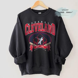 May include: Black sweatshirt with a baseball-themed graphic. The design features the word "Cleveland" in red, arched over a baseball player illustration and the text "EST. 1894". Two crossed baseball bats are below the illustration.