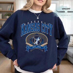 May include: Navy blue sweatshirt with a baseball design. The graphic reads "BASEBALL KANSAS CITY" in blue and white, with a baseball player illustration and "EST. 1969". The sweatshirt has a crew neck and long sleeves. The design is centered on the chest.