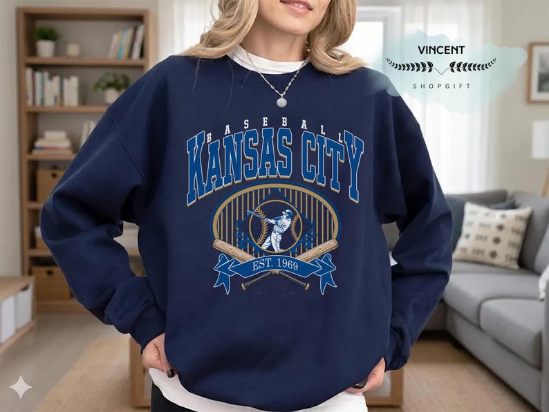 May include: Navy blue sweatshirt with a baseball design. The graphic reads "BASEBALL KANSAS CITY" in blue and white, with a baseball player illustration and "EST. 1969". The sweatshirt has a crew neck and long sleeves. The design is centered on the chest.