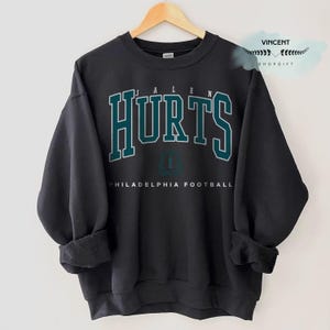 Puede incluir: Una sudadera negra con el nombre "HURTS" en letras de bloque turquesas con un contorno blanco. Debajo está el texto "PHILADELPHIA FOOTBALL". El número "1" está en el centro. La sudadera está colgada en una percha de madera.
