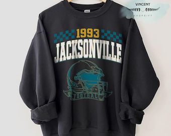 May include: Black sweatshirt with the text "1993 JACKSONVILLE" in white and gold, above a teal and black football helmet graphic. The sweatshirt is hanging on a wooden hanger.