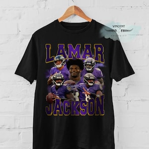 May include: Black t-shirt featuring a graphic of Lamar Jackson in a purple and gold design. The text "LAMAR JACKSON" is prominently displayed. The shirt is hanging on a wooden hanger.