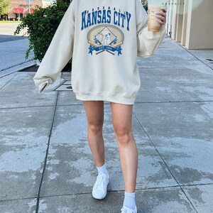May include: Cream-coloured sweatshirt featuring the text "Kansas City Baseball" in blue, with a baseball graphic. The sweatshirt has long sleeves and a crew neck. The image also shows white trainers and a drink.