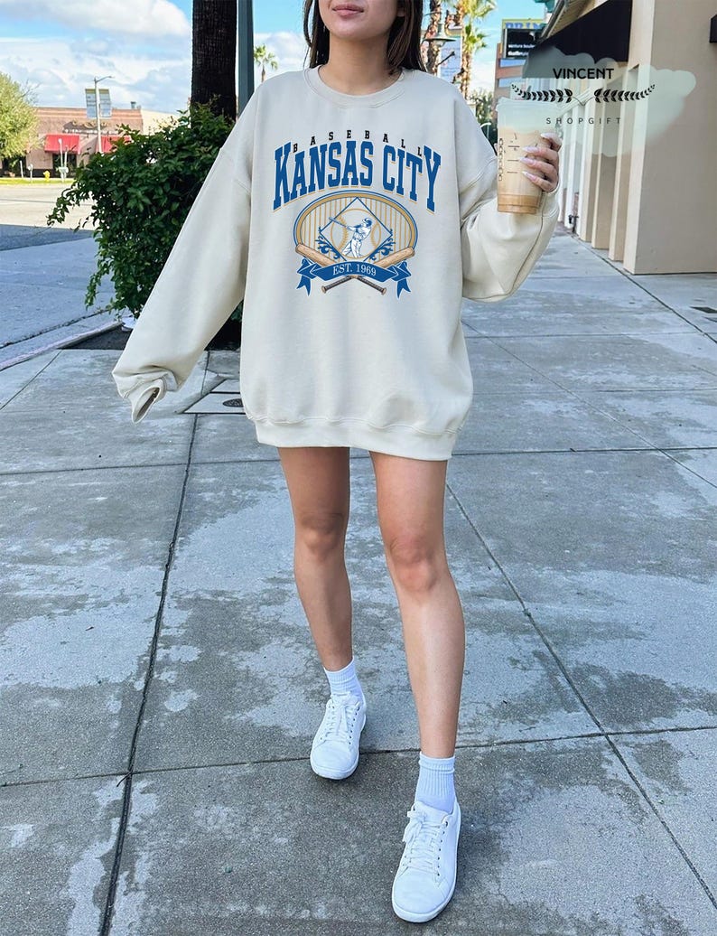 May include: Cream-coloured sweatshirt featuring the text "Kansas City Baseball" in blue, with a baseball graphic. The sweatshirt has long sleeves and a crew neck. The image also shows white trainers and a drink.