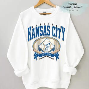 May include: White sweatshirt with a baseball graphic. The design includes the words "Kansas City" in blue, a baseball player illustration, crossed bats, and the text "EST. 1969". The sweatshirt is displayed on a wooden hanger.