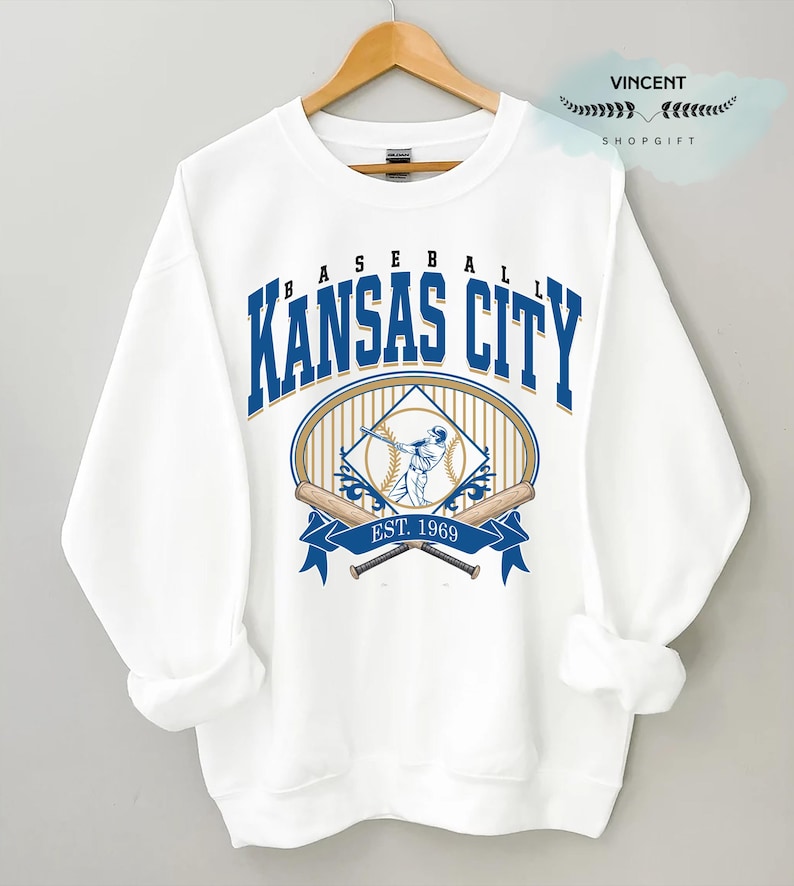 May include: White sweatshirt with a baseball graphic. The design includes the words "Kansas City" in blue, a baseball player illustration, crossed bats, and the text "EST. 1969". The sweatshirt is displayed on a wooden hanger.
