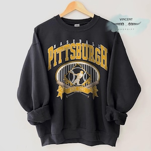 Puede incluir: Sudadera negra con la palabra "PITTSBURGH" en letras doradas, arqueada sobre un gráfico de béisbol. El gráfico incluye un jugador de béisbol, bates y el texto "EST. 1887". La sudadera está colgada en una percha de madera.