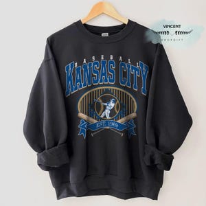 May include: Dark grey sweatshirt with "Kansas City Baseball" in blue and gold lettering. Features a baseball player graphic, crossed bats, and "EST. 1969". A vintage-style design, ideal for sports fans.