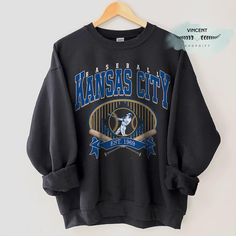 May include: Dark grey sweatshirt with "Kansas City Baseball" in blue and gold lettering. Features a baseball player graphic, crossed bats, and "EST. 1969". A vintage-style design, ideal for sports fans.