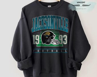 May include: Black sweatshirt featuring the text "JACKSONVILLE" in teal, with "1993" and a football helmet graphic. The word "FOOTBALL" is printed at the bottom. The sweatshirt is hanging on a wooden hanger.