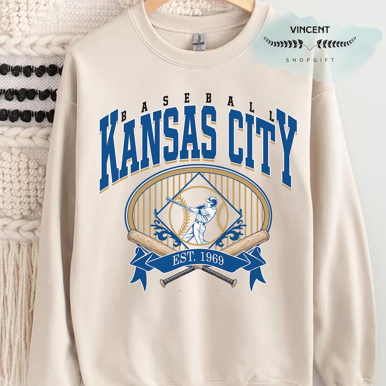 May include: Cream-coloured sweatshirt with a blue and gold baseball graphic. The design features the words "Kansas City", a baseball player, baseball bats, and the text "EST. 1969". The sweatshirt is a neutral colour.