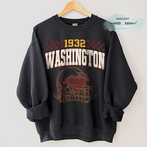 May include: Black sweatshirt with the text "1932 WASHINGTON FOOTBALL" in white and gold lettering. The design includes a football helmet in burgundy and gold. The sweatshirt is hanging on a wooden hanger.