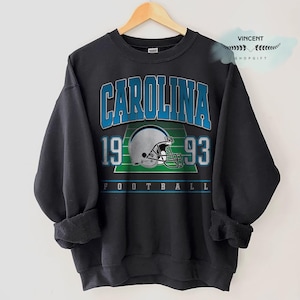 Puede incluir: Sudadera negra con la palabra "CAROLINA" en letras azules en bloque sobre los números "19" y "93". Un gráfico de casco de fútbol está centrado en un campo verde. La palabra "FOOTBALL" está impresa en la parte inferior.