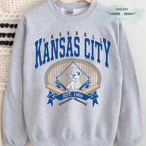 May include: A light grey sweatshirt with a baseball design. The design features the words "BASEBALL KANSAS CITY" in blue and gold, a baseball player graphic, crossed bats, and the text "EST. 1969".
