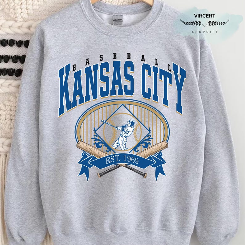 May include: A light grey sweatshirt with a baseball design. The design features the words "BASEBALL KANSAS CITY" in blue and gold, a baseball player graphic, crossed bats, and the text "EST. 1969".