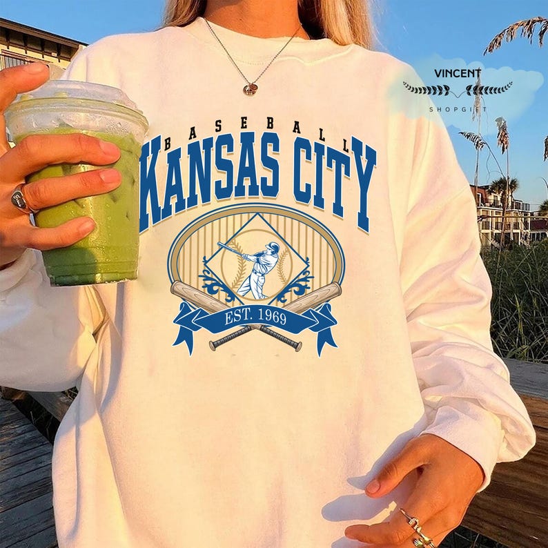 May include: White long-sleeved sweatshirt with a blue and white baseball graphic. The graphic says "Kansas City" above a baseball player illustration and "Est. 1969". The sweatshirt is a casual style.