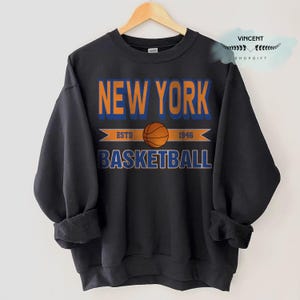 May include: Black sweatshirt with the words "NEW YORK BASKETBALL" in orange and blue lettering. A basketball graphic is centered below the words "NEW YORK". The sweatshirt also has the text "ESTD 1946". The sweatshirt is hanging on a wooden hanger.