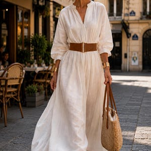 May include: A woman in a long, white linen dress with a brown belt and a straw tote bag. The dress has long sleeves and a V-neck. She is wearing gold bracelets and sunglasses, walking on a cobbled street.