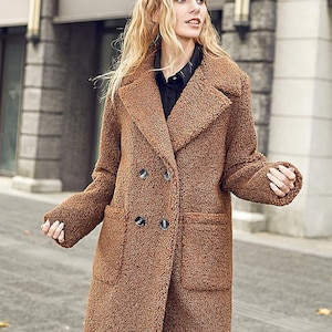 Könnte beinhalten: Ein brauner Teddy-Mantel mit doppelter Knopfleiste, breiten Revers und zwei Vordertaschen. Der Mantel hat dunkle Knöpfe und ein strukturiertes, weiches Aussehen. Das Model trägt ein schwarzes Hemd. Der Mantel ist knielang.