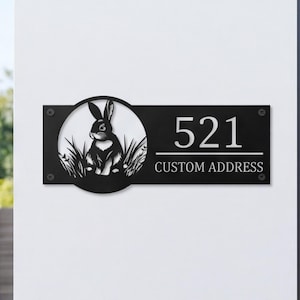 May include: A rectangular black metal address sign featuring a circular cutout on the left side with a silhouette of a rabbit sitting in grass. The right side displays the number 521 above the text CUSTOM ADDRESS in a white serif font.