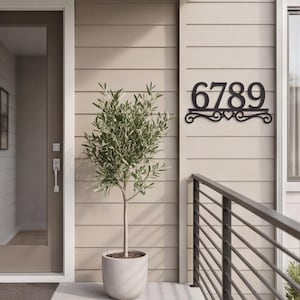 May include: A black metal house number sign displaying the digits 6789 in a bold serif font. The sign features a decorative scrollwork base with a central heart shape, mounted horizontally on a light beige horizontal siding wall surface.