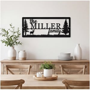 May include: A rectangular black metal wall sign featuring a silhouette of two pine trees and a deer. The text reads the MILLER family in a combination of script and serif fonts. The sign is mounted horizontally with a thin border frame.