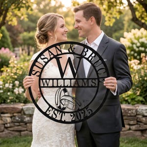 Puede incluir: Un letrero de boda de metal negro con los nombres "Susan & Bruce" y "Williams" y la fecha "EST. 2025". El letrero también incluye un gráfico de dos anillos de boda. El letrero lo sostiene una pareja en un jardín.