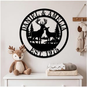 May include: A circular black metal wall sign featuring a silhouette of a stag and a doe standing in a forest landscape. The text DANIEL & AMELIA is arched across the top, and EST. 1975 is displayed along the bottom edge of the circular frame.