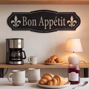 May include: A black rectangular wall sign with decorative curved edges featuring the white text Bon Appétit and a fleur-de-lis symbol on each side. The sign is mounted on a wall above a wooden shelf containing a coffee maker and a lamp.
