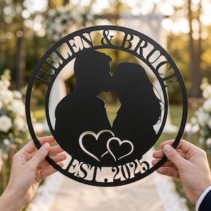 May include: Black metal wedding sign featuring a kissing couple silhouette, two hearts, and the text "HELEN & BRUCE EST. 2025" in a circular design. The sign is held up by two hands.