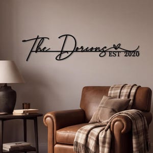May include: A black metal wall sign featuring the cursive text The Dorions and the text EST 2020, accented with a small heart symbol. The sign is mounted on a plain wall above a brown leather armchair and a side table with a lamp and books.