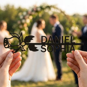 Puede incluir: Adorno para tarta de boda de madera negra cortada con láser con un diseño de rosa y los nombres "DANIEL" y "SOPHIA". Los nombres están en negrita, con una fuente sans-serif. El adorno se sostiene frente a una ceremonia de boda.