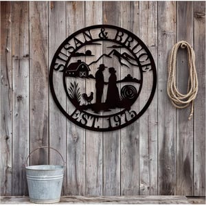 Puede incluir: Un letrero de pared circular de metal negro con la silueta de una pareja en un paisaje rural con un granero, montañas y un gallo. El texto SUSAN & BRUCE está arqueado en la parte superior y EST. 1975 está arqueado en la parte inferior.