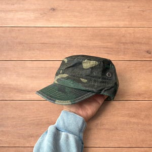 Puede incluir: Una gorra de estilo militar con un patrón de camuflaje en verde oscuro, marrón y negro. El accesorio presenta una parte superior plana, una visera corta y curva, y un pequeño ojal metálico de color plateado en el lateral para ventilación.