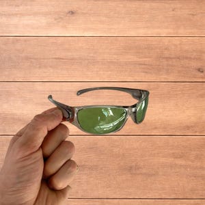 May include: Sport sunglasses with a grey frame and green lenses. The sunglasses have a sleek, modern design, suitable for outdoor activities. The lenses are a vibrant green colour, offering protection from the sun.