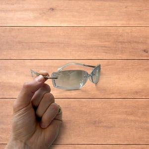 May include: Rimless sunglasses with a light blue tint. The rectangular lenses are held by a hand. The frame and arms are a clear, light blue colour. The sunglasses are a stylish accessory.