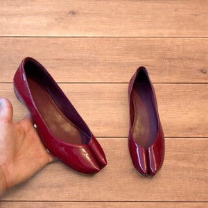 Pode incluir: Um par de sapatos rasos de verniz vermelho escuro com um design distinto de biqueira dividida. Os sapatos têm um acabamento brilhante, um peito do pé baixo e um forro interior liso. A construção da biqueira separa o dedo grande do pé.