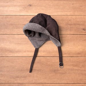 May include: A dark brown corduroy hat featuring a textured, light grey fleece lining. The hat includes two side flaps that extend downwards, with a thin corduroy strap attached to one side, finished with a circular metal ring for fastening.
