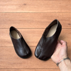 Op de afbeelding: Een paar donkerbruine leren schoenen met gespleten teen, ook wel Tabi-schoenen genoemd, met een lage hak en een splitsing voor de grote teen. De schoenen worden gepresenteerd op een houten oppervlak.
