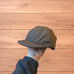 Può includere: Un cappellino da baseball grigio con visiera curva. Il cappellino è realizzato in un tessuto resistente e ha un design classico e discreto. Il cappellino è tenuto da una mano.