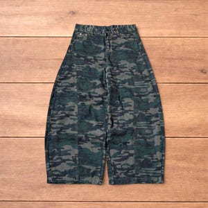 May include: Camouflage-patterned pants in shades of green and gray. The pants have a wide-leg design and a classic five-pocket style. The fabric appears to be denim or a similar durable material. The pants are laid flat on a wooden surface.