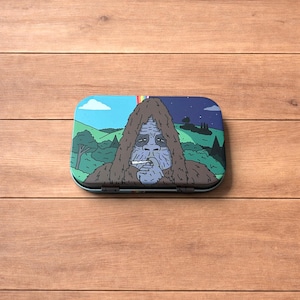 Peut inclure: Une boîte métallique rectangulaire avec une illustration colorée d'un Bigfoot fumant une cigarette. Le fond représente un paysage avec un arc-en-ciel, des arbres et un ciel nocturne. La boîte est sur une surface en bois.